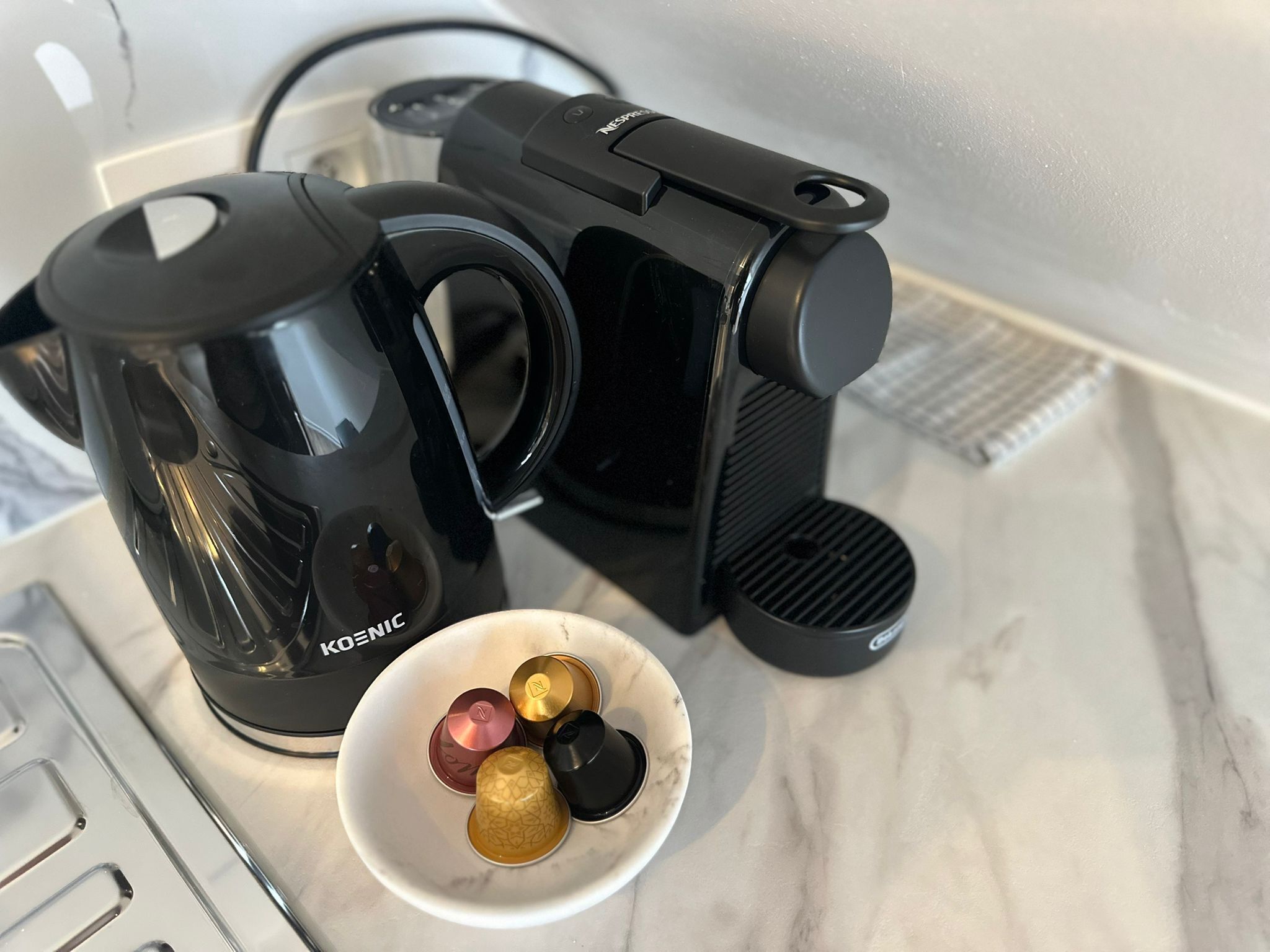 Una cafetera, una jarra de vidrio y un bol con cápsulas de café sobre una encimera de mármol.