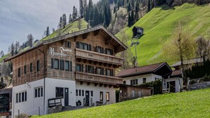 Een tweelaags landhuis met een houten gevel en een balkon, gelegen voor een achtergrond van groene heuvels en een heldere lucht.