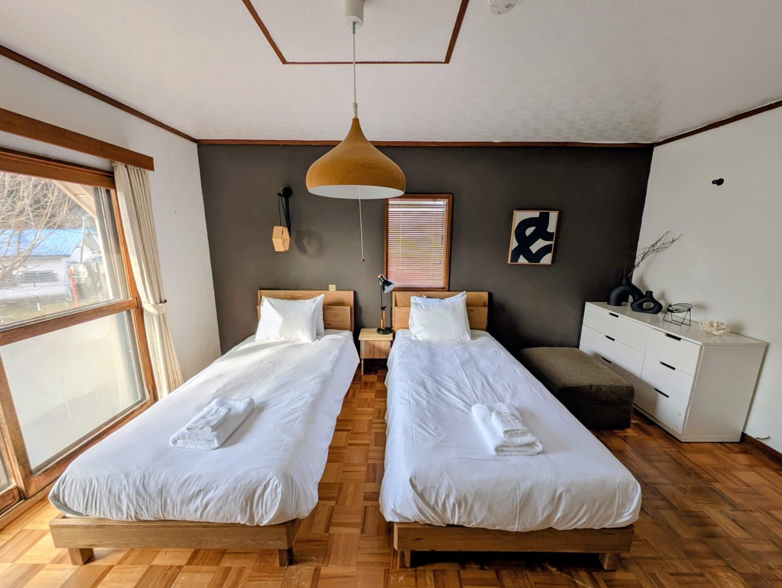 Quarto com duas camas, piso de madeira, uma janela grande, luminária pendente e cômoda branca.