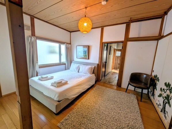 A bedroom with a bed, a wooden ceiling, a window with curtains, a painting on the wall, and a chair.