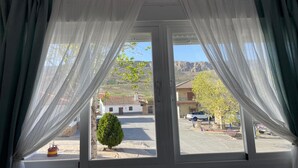 Ein Zimmer mit großem Fenster, weißen Vorhängen und Blick auf eine Straße mit Gebäuden und einem Berg im Hintergrund.