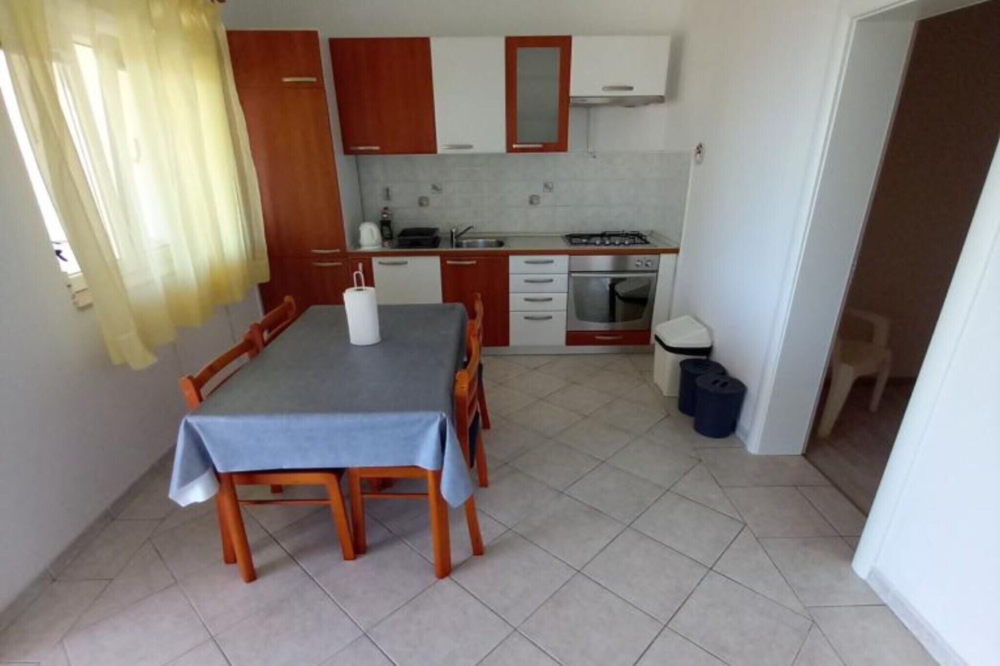 A compact kitchen with a dining table, wooden cabinets, a stove, and a trash bin.