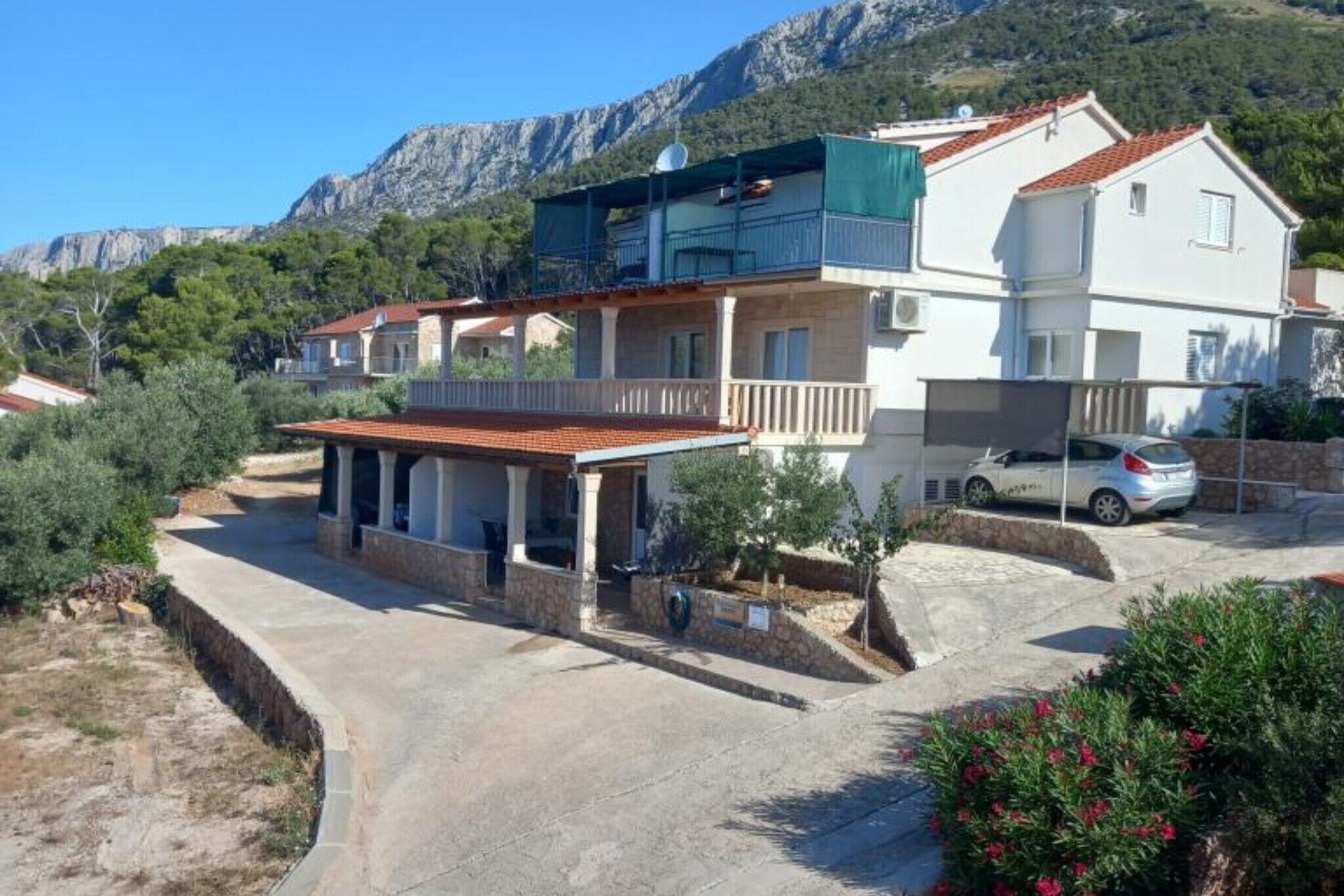 A two-story house with a balcony, a car parked in front, and a mountainous backdrop.