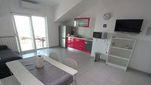 A modern kitchen with red cabinets, a white countertop, and a black TV mounted on the wall. There is a dining area with a white table and chairs, and a large window providing natural light. - One Bedroom Apartment with Balcony and Sea View (Sveta Nedjelja)
