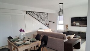 A modern living room with a sofa, dining table, and chairs. There is a staircase with a metal railing, a flat-screen TV mounted on the wall, and a vase with flowers on the table. - Elegant Villa Ilma with Pool - Villa With Pool (Vodice)