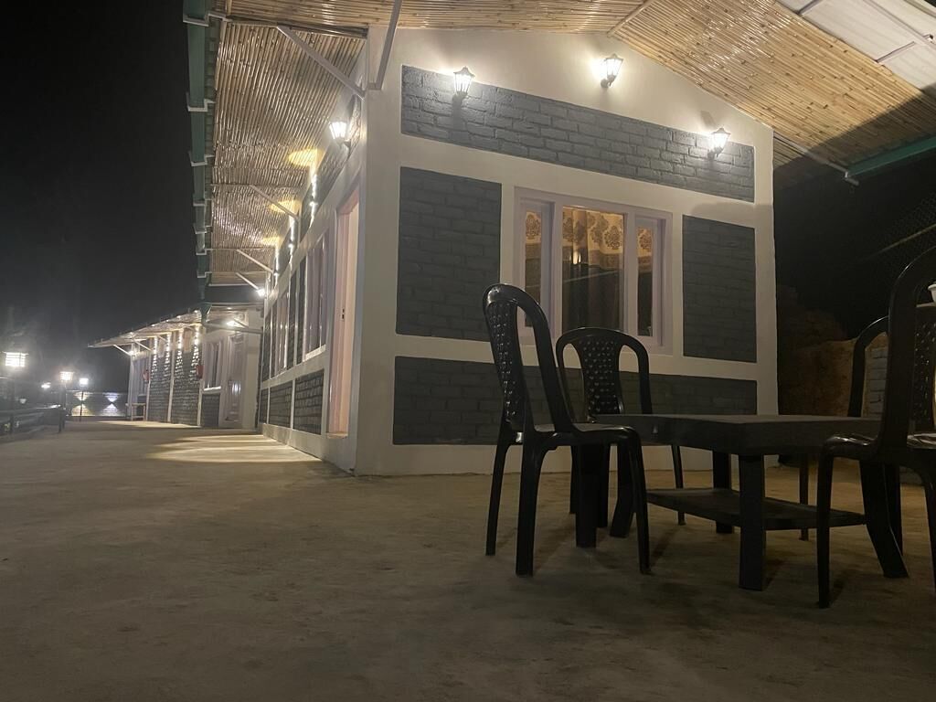 A small, single-story building with a flat roof and a brick facade. There are two chairs and a table in front of the building, and the area is illuminated by overhead lights.