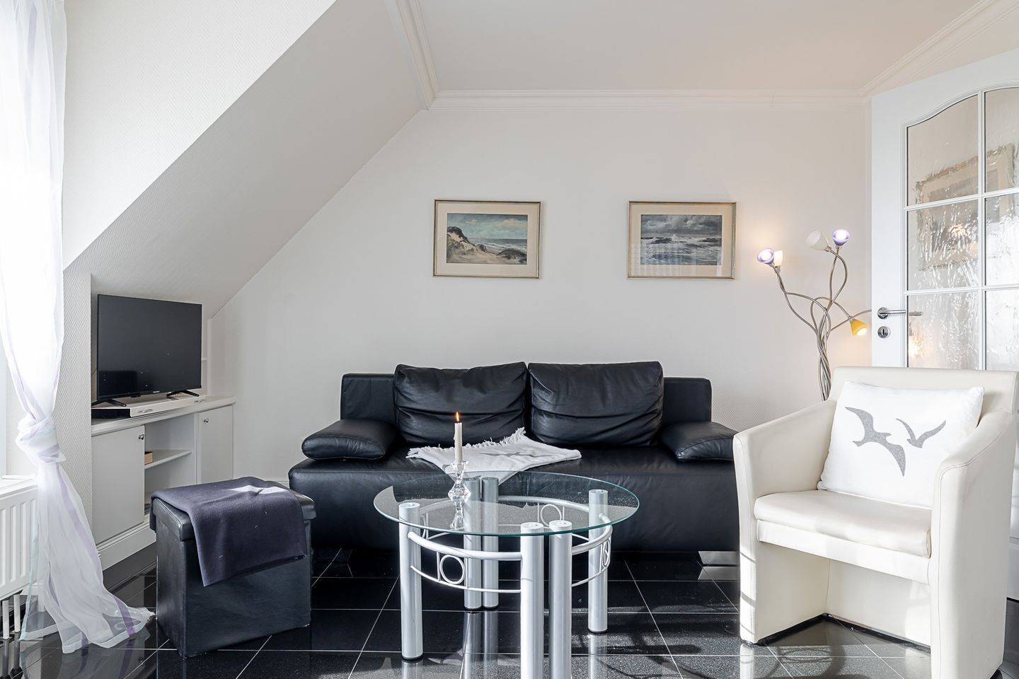 Una sala de estar moderna con un sofá de cuero negro, una mesa de centro de vidrio, un sillón blanco y dos cuadros en la pared.