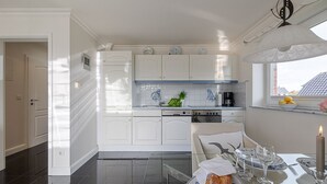A modern kitchen with white cabinetry, a dining area with a table set for two, and a window with a view of buildings. - Sand - Lust auf Sylt - Sand vacation apartment for 2 people (Sylt-Ost)