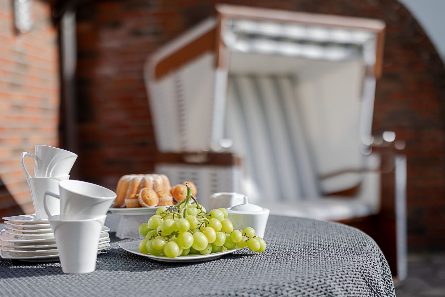 A table with white cups, saucers, a plate of green grapes, and a basket of pastries.