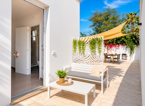 Une terrasse ensoleillée avec une table blanche, un banc rembourré et un paravent décoratif agrémenté de plantes suspendues.