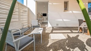 Un espace extérieur aménagé avec des chaises et une table en métal, un mur blanc avec une fenêtre et une porte donnant sur un espace intérieur.