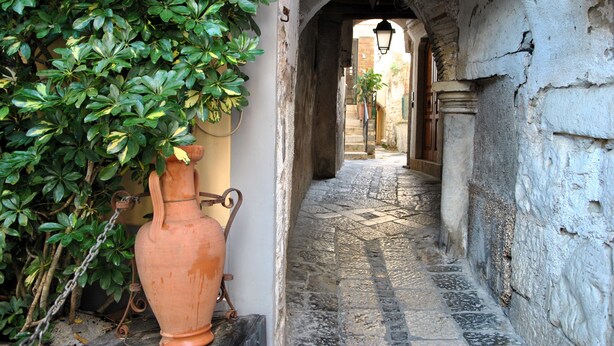 Un estrecho callejón empedrado con una maceta rústica y follaje a la izquierda, que conduce a un edificio con un arco de piedra y una lámpara colgante.