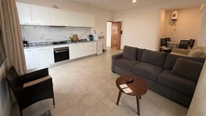 Un salon moderne avec un canapé gris, une petite table basse ronde et un fauteuil en osier. On aperçoit, au fond, une cuisine avec des meubles blancs et un pare-feu en marbre.