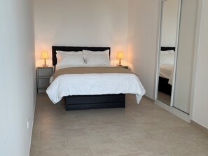 A neatly made bed with white bedding and a dark headboard, flanked by two bedside tables with lamps, in a room with a tiled floor and a large mirror on one wall.