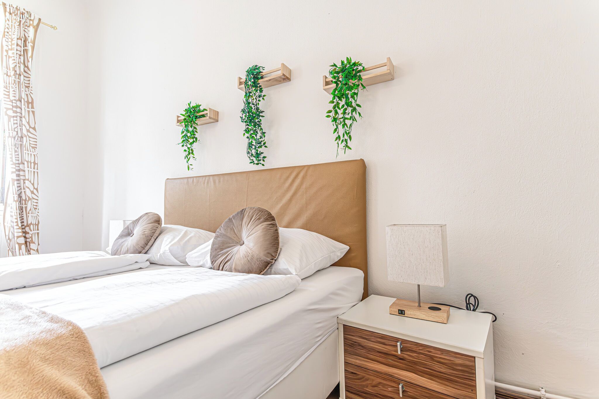 A neatly made bed with pillows, a bedside table with a lamp, and three hanging plants on the wall.