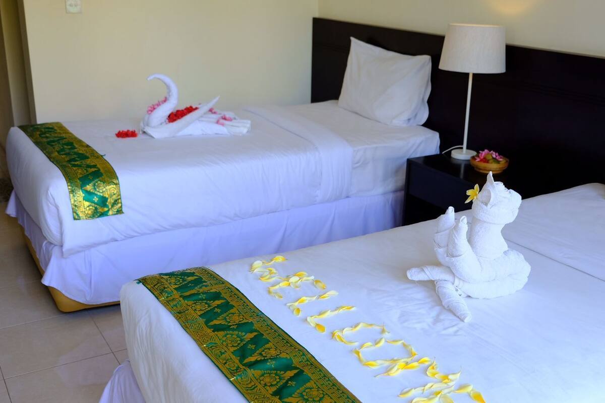 Two hotel beds with white linens, one with a green and gold headboard, and a towel sculpture on the right bed.