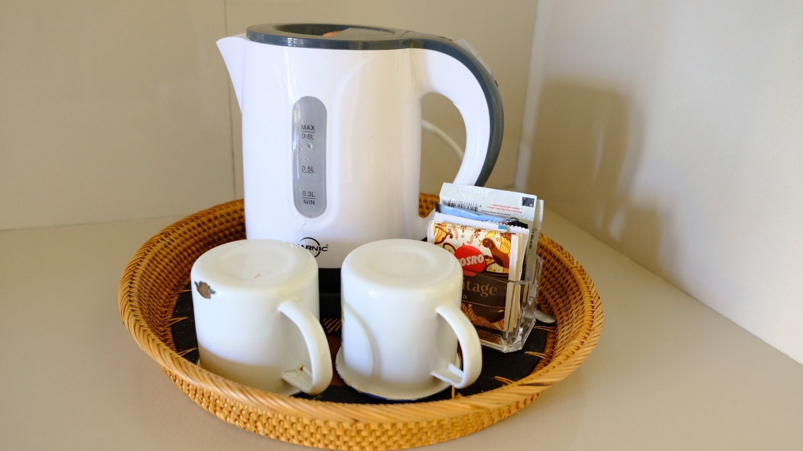 A white electric kettle, two white mugs, and a coffee sachet on a woven tray.
