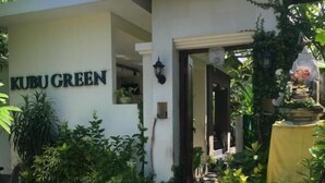 Egy épület, amelyen egy „KUBU GREEN” feliratú tábla látható, és egy ajtó vezet a belső térbe.