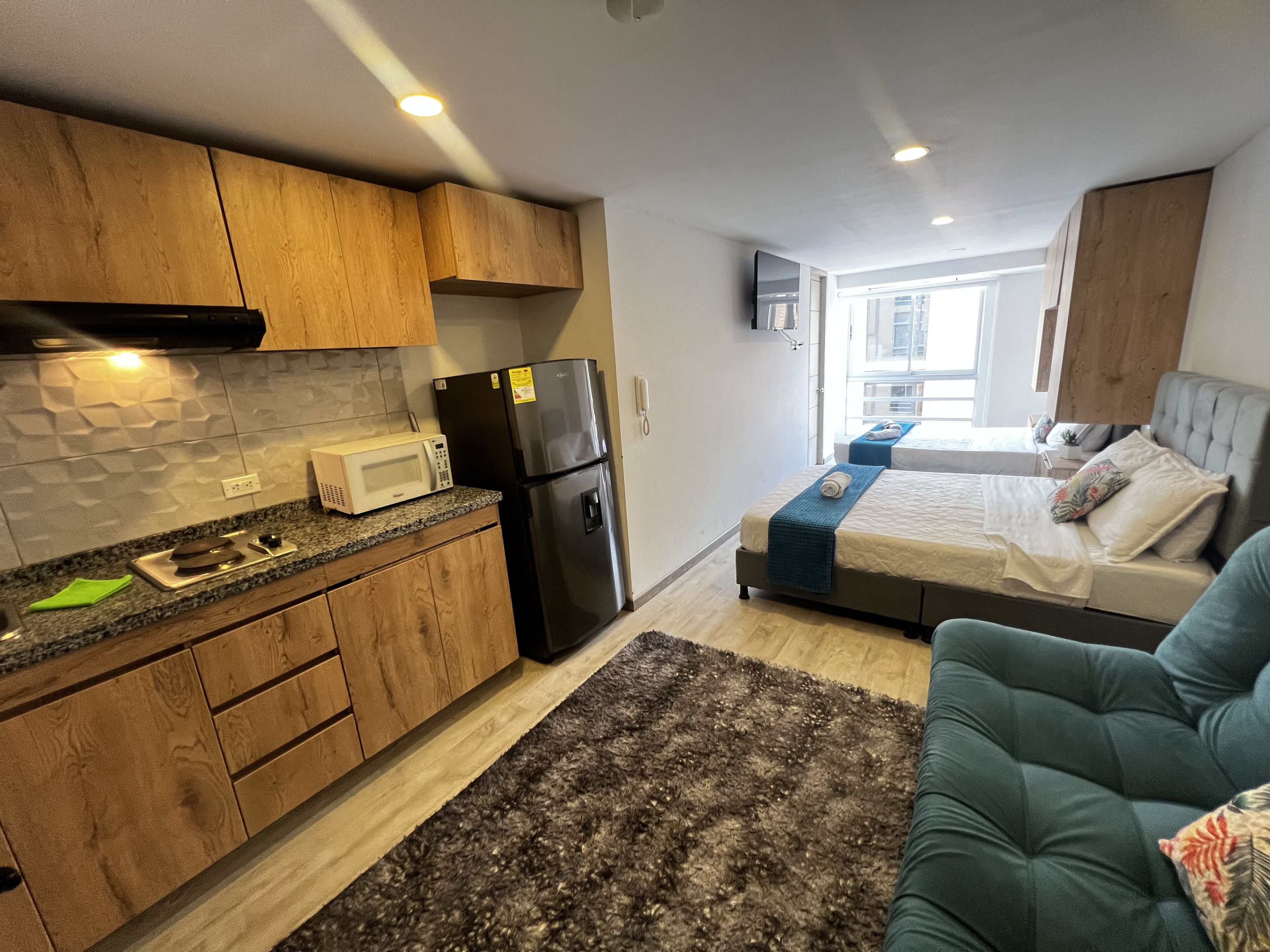 Habitació d'hotel compacta amb petita cuina, un llit amb llençol blau i un sofà gris.