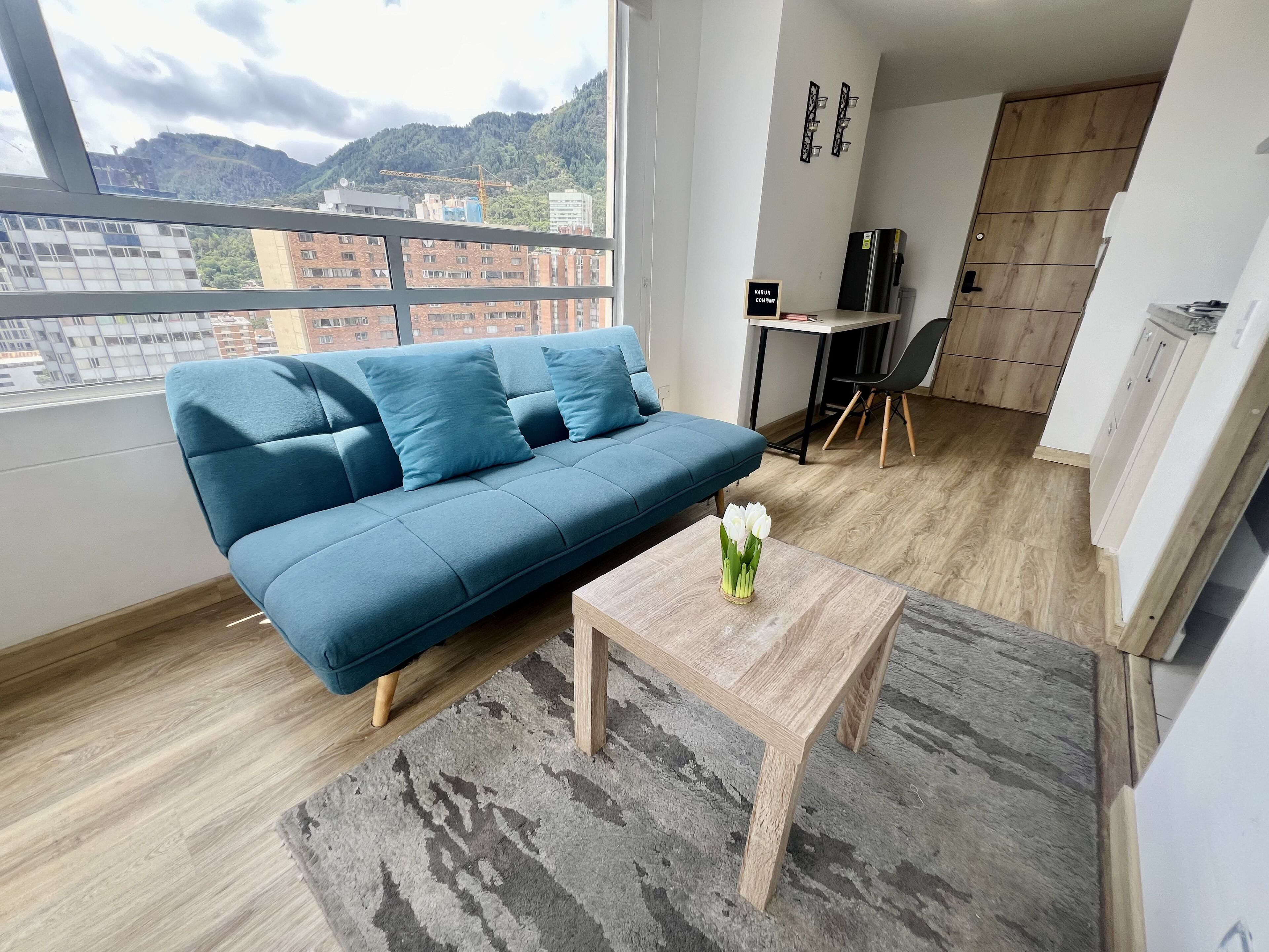 A modern living room with a blue sofa, a small wooden table with a vase of white flowers, and a view of a cityscape with mountains in the background.