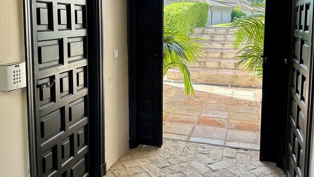 A hallway with a stone floor leading to an open door revealing a courtyard with a stone pathway, greenery, and a building in the background. - PATIO ROOM SOTOGRANDE (sotogrande)