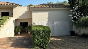 A building with a tiled roof, a white garage door, and a well-maintained garden with a tall tree and potted plants. - PATIO ROOM SOTOGRANDE (sotogrande)
