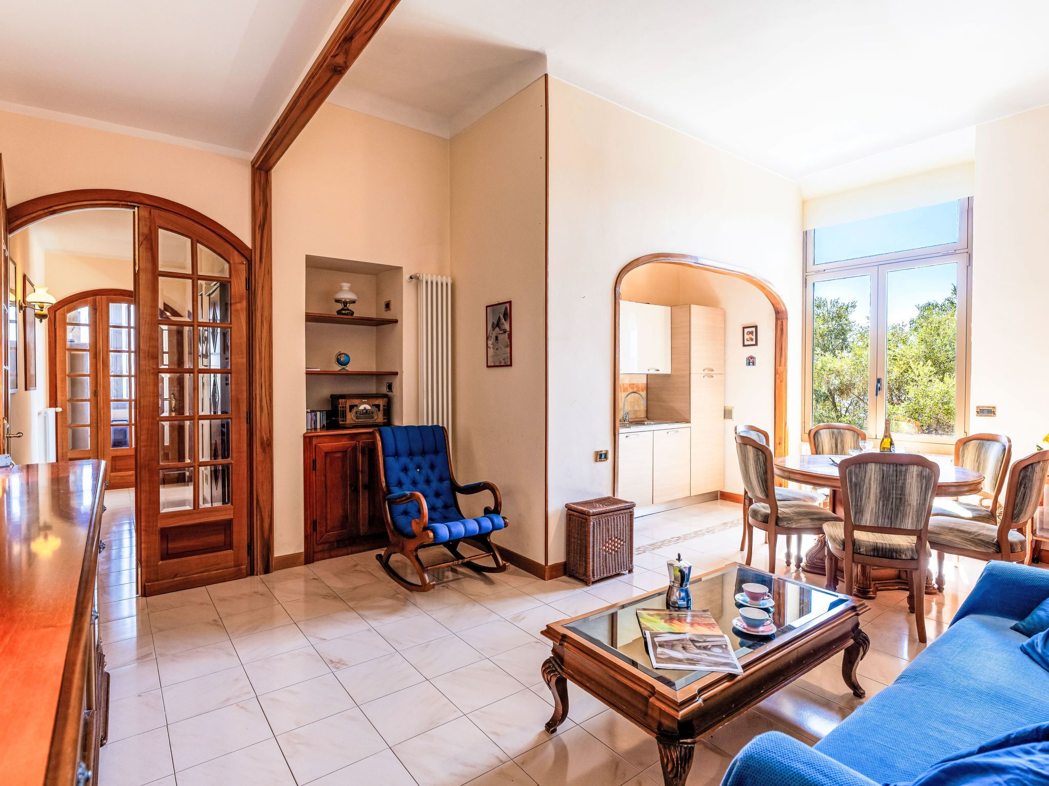 A spacious living area with a wooden-framed door, a blue armchair, a glass-top coffee table, and a dining area with a view of the outdoors.