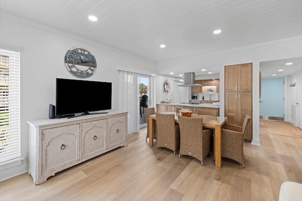 A modern kitchen with white cabinets, a dining area with wicker chairs, a flat-screen TV on a white cabinet, and a wall clock.
