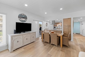 A modern kitchen with white cabinets, a dining area with wicker chairs, a flat-screen TV on a white cabinet, and a wall clock.