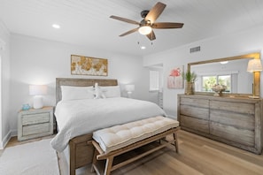 A bedroom with a bed, bedside tables, a bench, a ceiling fan, a dresser, and artwork on the walls.