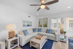 A living room with a sofa, coffee table, and side tables, a ceiling fan, and artwork on the walls.