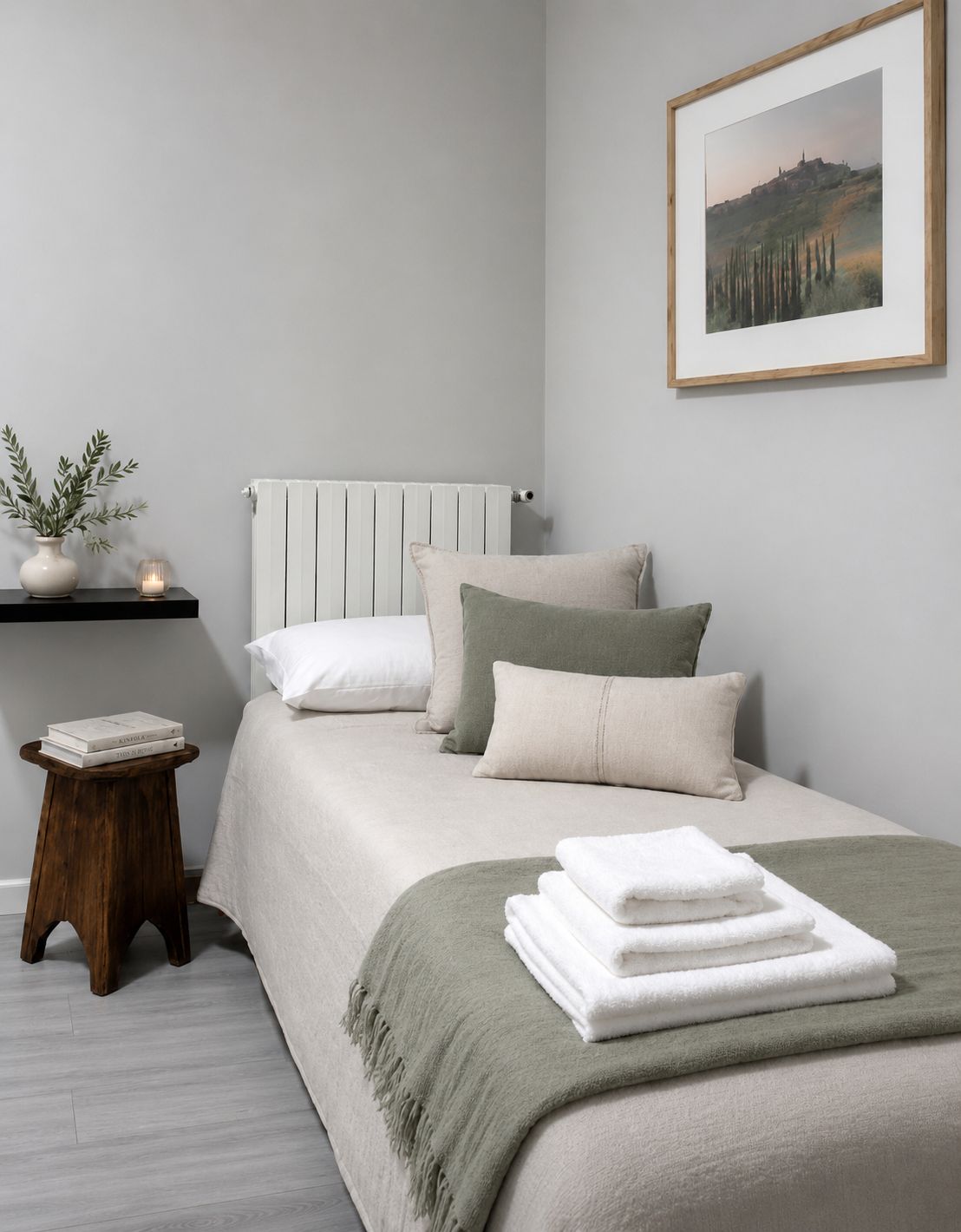 Une chambre à coucher bien rangée, avec un lit, des oreillers, une table de chevet agrémentée d’une plante, un tableau encadré au mur et un radiateur.
