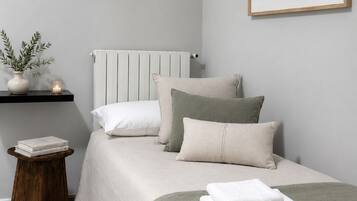 Une chambre à coucher bien rangée, avec un lit, des oreillers, une table de chevet agrémentée d’une plante, un tableau encadré au mur et un radiateur.