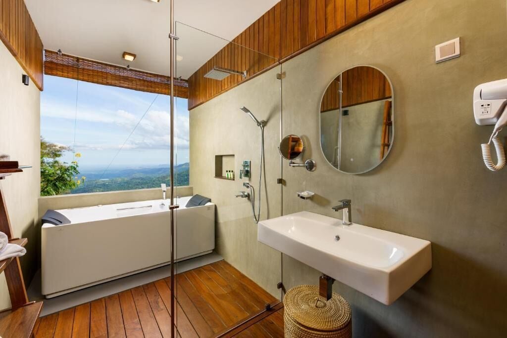 A modern bathroom with a large bathtub, a sink, and a shower. There is a view of the outdoors through a large window.
