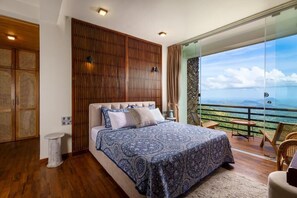 Een slaapkamer met een groot bed, een houten hoofdbord, een klein nachtkastje en uitzicht op de oceaan door een glazen deur.