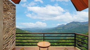 A balcony with a wooden table and two wicker chairs, overlooking a mountainous landscape.