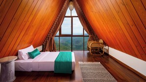 A cozy attic bedroom with a wooden sloped ceiling, a large window offering a view of the landscape, a bed with a green throw, and a wicker chair.