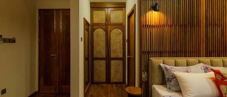 A room with a wooden floor, a bed with floral bedding, a wall-mounted lamp, and a wooden door with a woven panel.