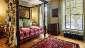 A four-poster bed with a red and black patterned bedspread, a large red and black area rug, and a view of the street through a large window with green curtains.