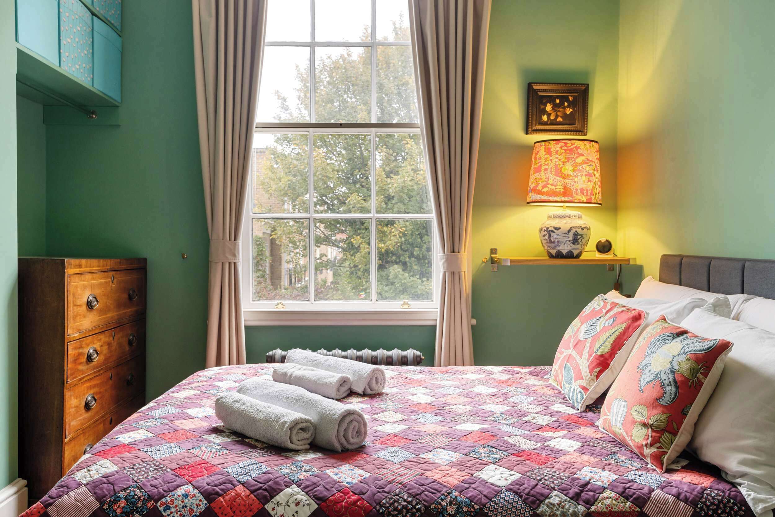 Une chambre à coucher avec une couvre-lit matelassé, une commode en bois, une fenêtre avec des rideaux et une lampe de chevet.