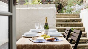 An outdoor dining setup with a table set for two, featuring plates, glasses, a bottle of wine, cheese, and grapes.