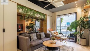 Een moderne woonkamer met een grijze bank, een ronde salontafel en een plafondventilator. Er is een groot raam met uitzicht op de stad.