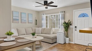 A modern living room with a sofa, a dining table with placemats, a ceiling fan, and a large window with a view of a staircase. - 2BR Beach Getaway w/ Patio, AC, WiFi, W&D, Smart TV's & Steps to the Sand (Newport Beach)