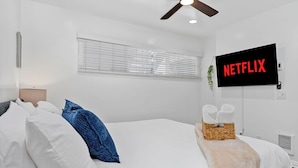 A hotel room with a bed, a ceiling fan, a TV mounted on the wall, and a window with blinds. - 2BR Beach Getaway w/ Patio, AC, WiFi, W&D, Smart TV's & Steps to the Sand (Newport Beach)
