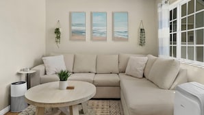 A modern living room with a beige sectional sofa, a round wooden coffee table, and three framed beach paintings on the wall. - 2BR Beach Getaway w/ Patio, AC, WiFi, W&D, Smart TV's & Steps to the Sand (Newport Beach)