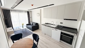 A modern kitchen with white cabinets, a black countertop, and a built-in oven. There is a dining area with a wooden table and black chairs. - Moon City (Antalya)