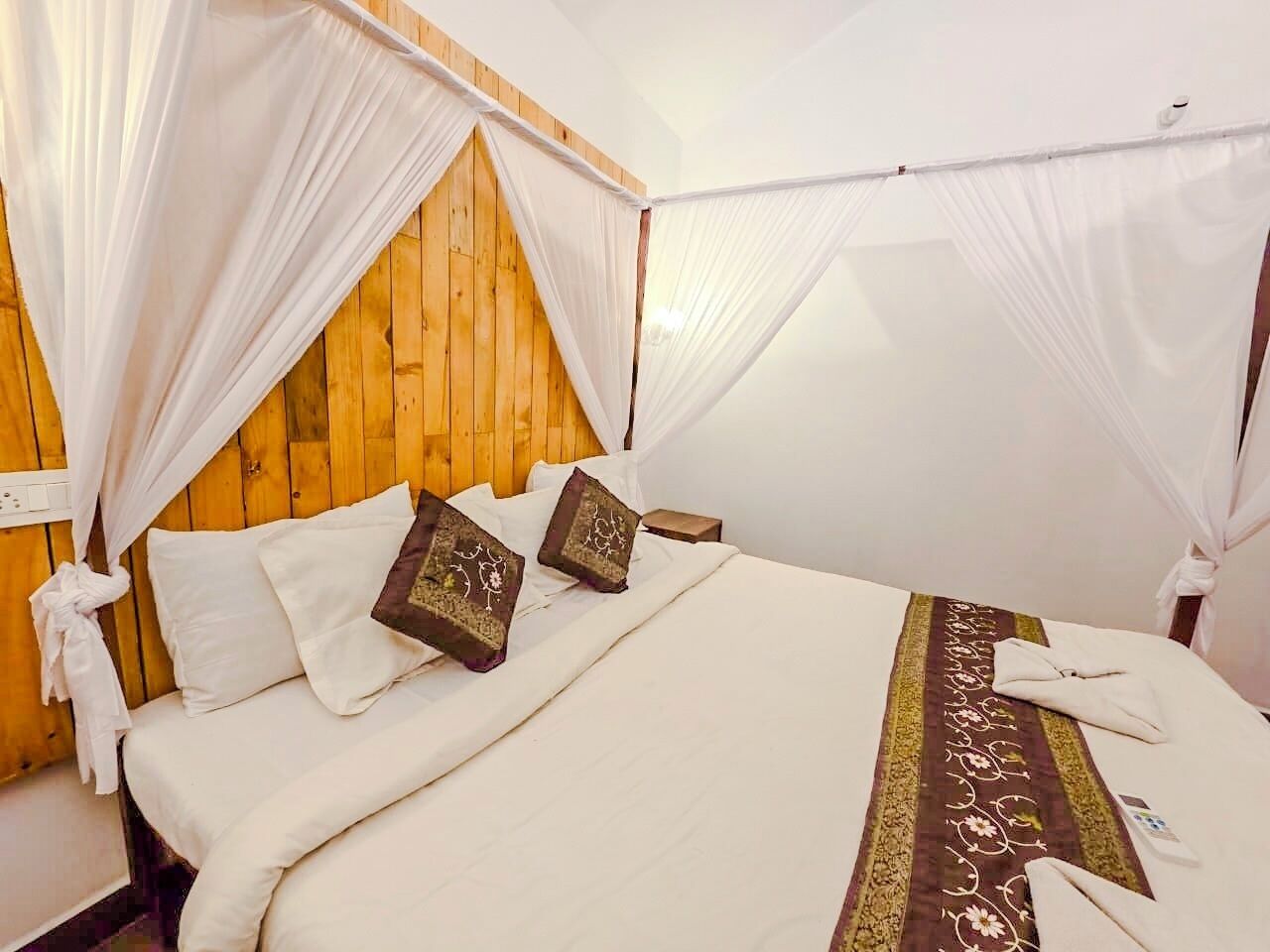 Una cama con ropa de cama blanca, dos almohadas decorativas, una manta marrón estampada y un dosel.