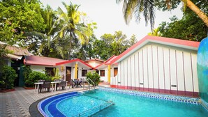 A swimming pool with a tiled edge, surrounded by a patio area with tables and chairs, and a building with a red roof and white walls. - Harbor Court Vagator (Vagator)