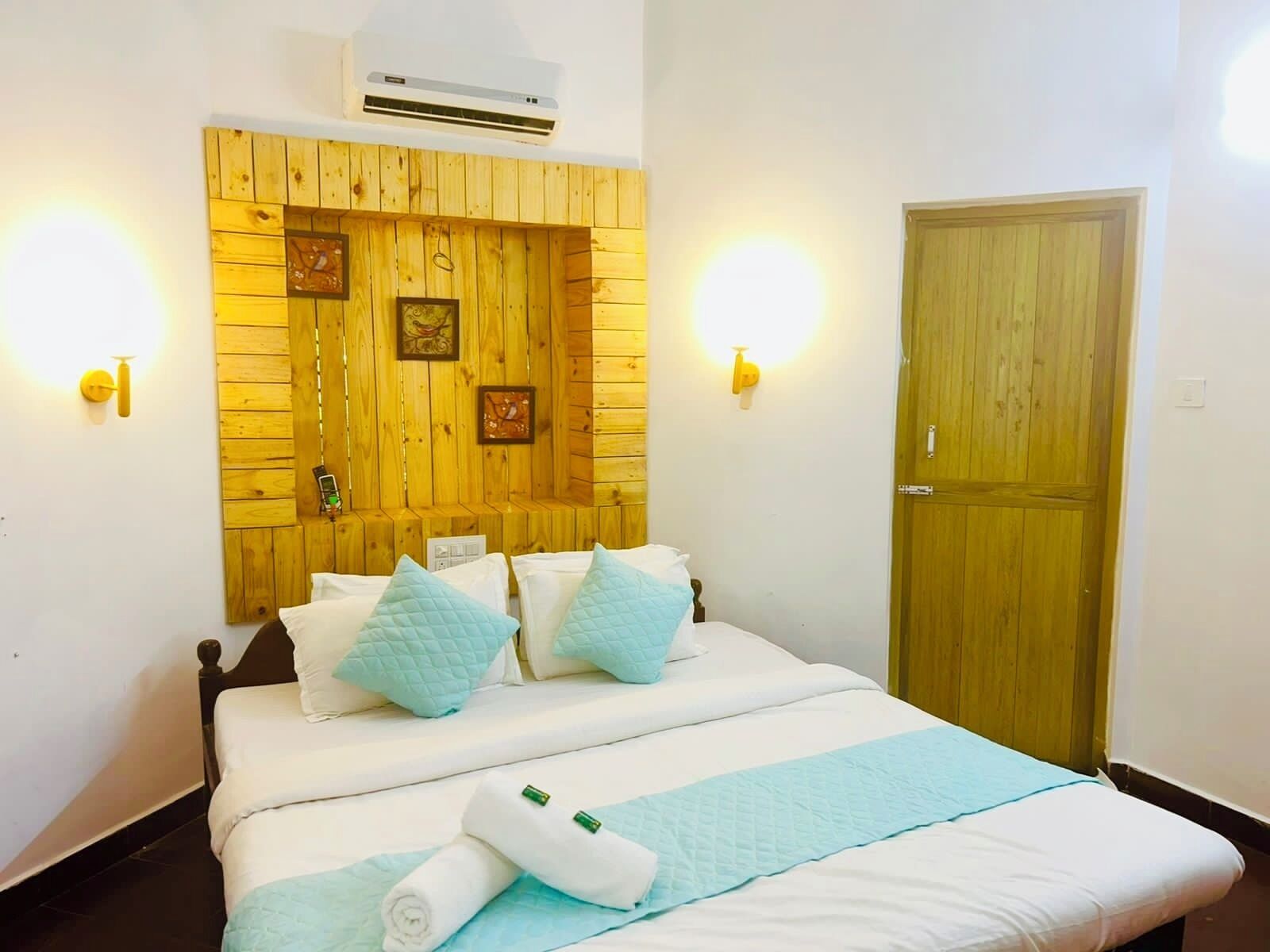 A hotel room with a wooden headboard, two wall-mounted lights, a door, and a bed with white bedding and blue pillows.