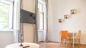Une pièce avec une table ronde, deux chaises, un téléviseur fixé au mur et une fenêtre avec des rideaux.